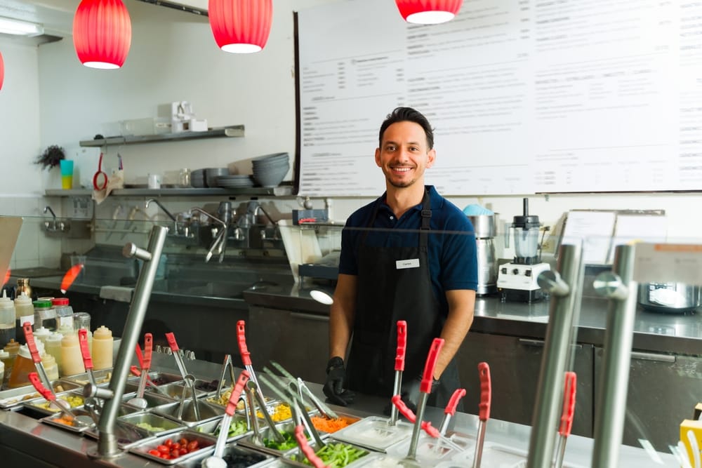 male worker wearing black apron and gloves standing behind salad bar counter offering healthy fresh food options